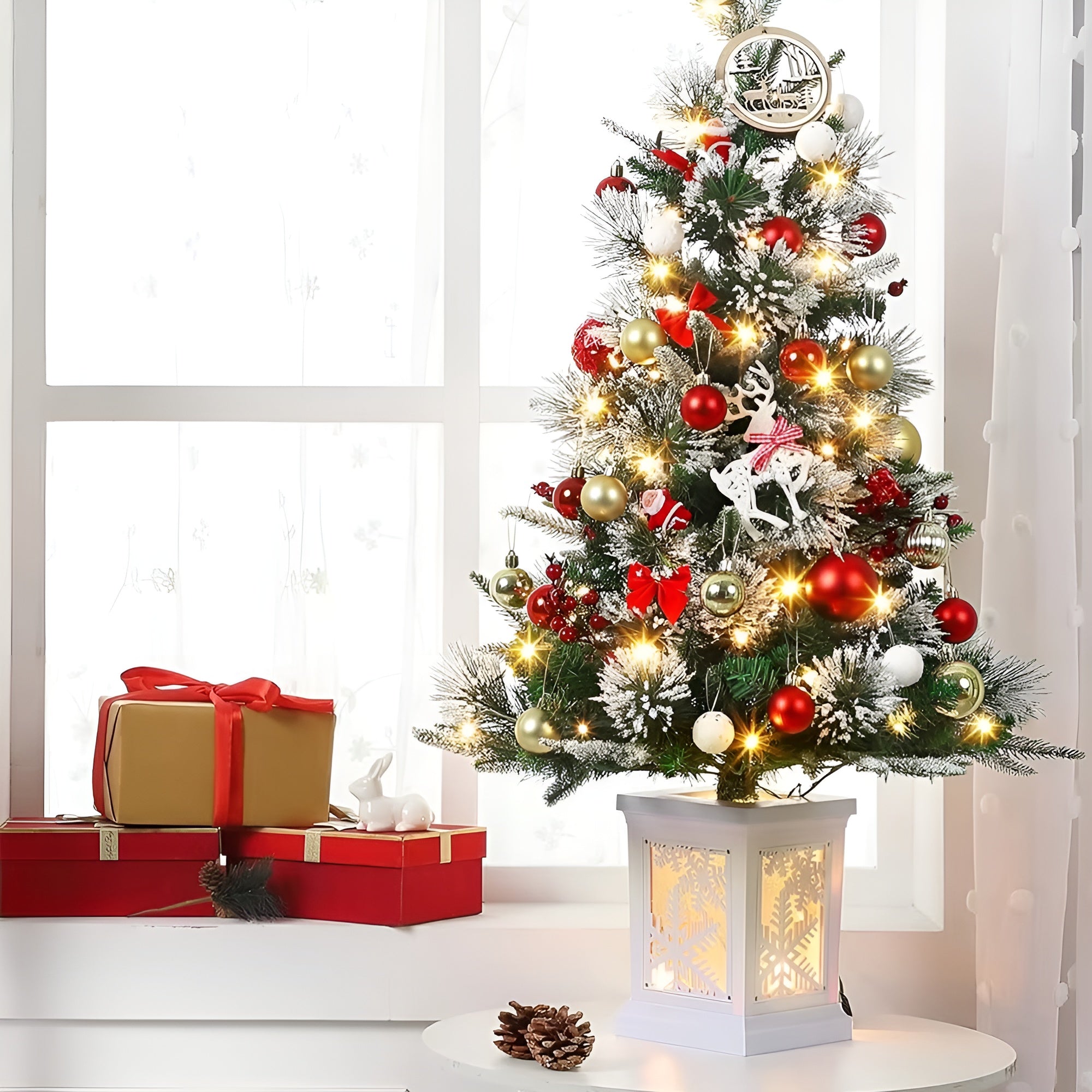 Decorated Christmas tree with ornaments and lights on a white surface, with gift boxes and a lantern in the background.