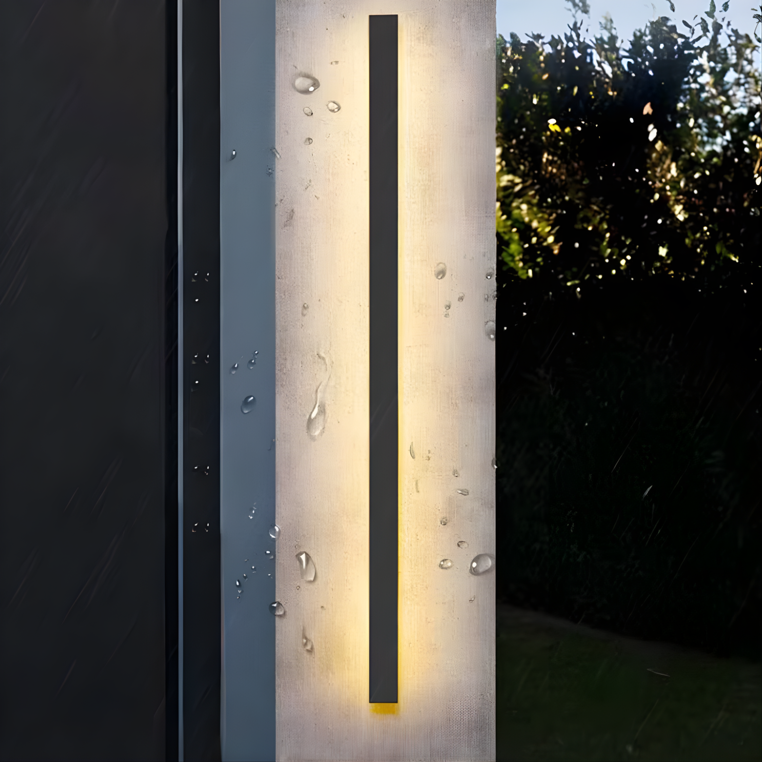 Modern outdoor wall light fixture on a concrete pillar with raindrops, surrounded by greenery.