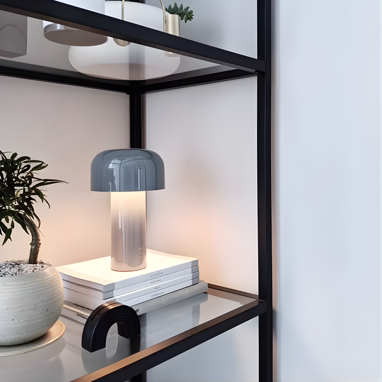 Modern table lamp on a stack of books with a plant and decorative items on a shelf.